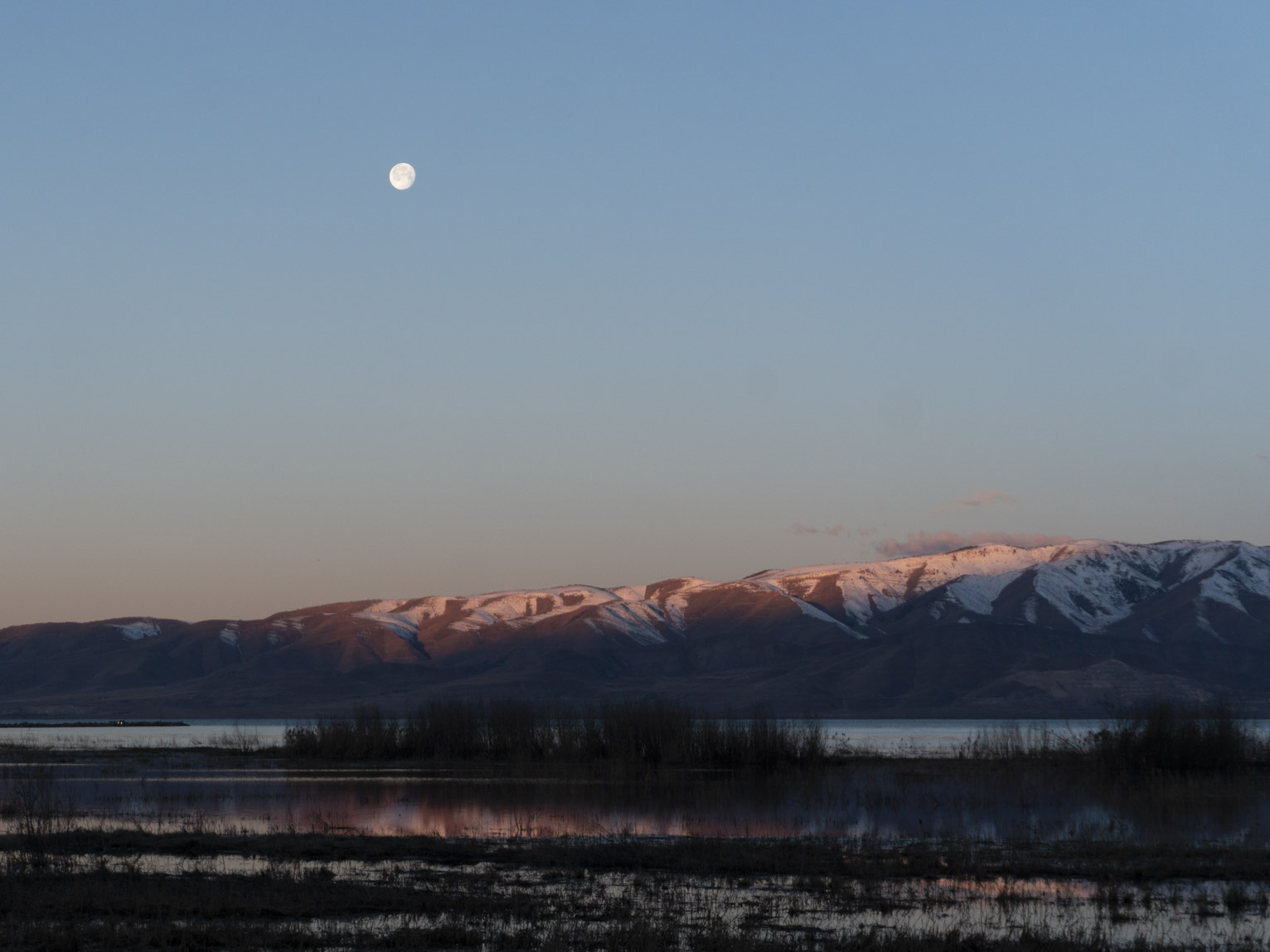 Snowy mountains across the lake catch the first days sun, the moon above